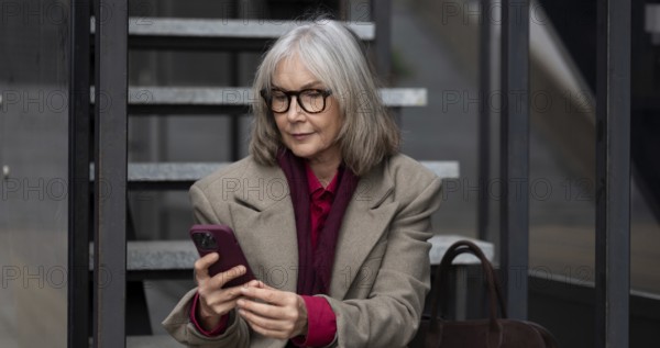 A middle-aged businesswoman dressed in a stylish coat and glasses is engaged with her smartphone while seated on outdoor steps. She appears focused and relaxed