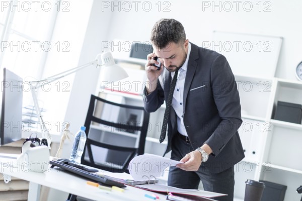 A man is standing near a table in the office, talking on the phone and flipping through documents