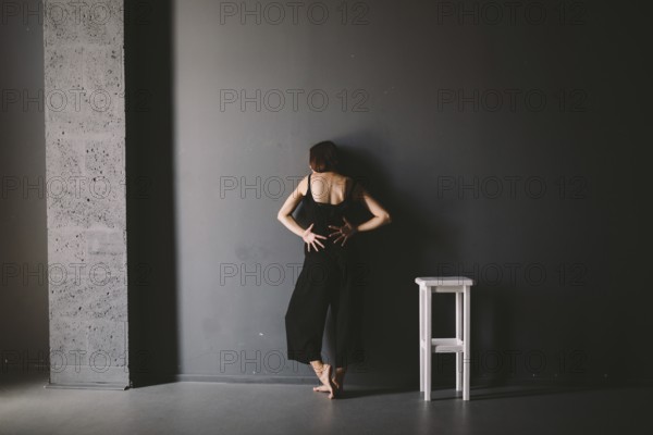 Social theme of female loneliness is pain suffering. Abastration of male domestic violence. Young beautiful Caucasian woman in black clothes, strange pose, white chair big dark room. Fear and despair