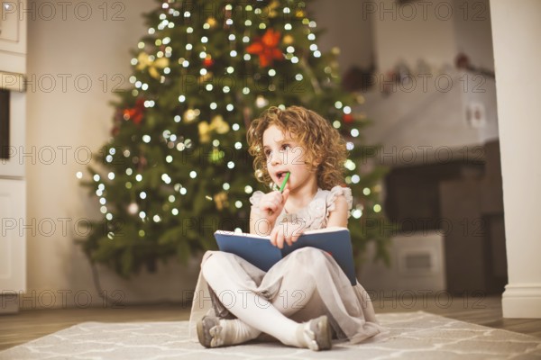 Sweet dreamy little girl with curly hair, wearing grey dress, sitting in front of decorated Christmas tree, with her legs crossed. Girl thinking about letter to Santa, holding notebook and pencil