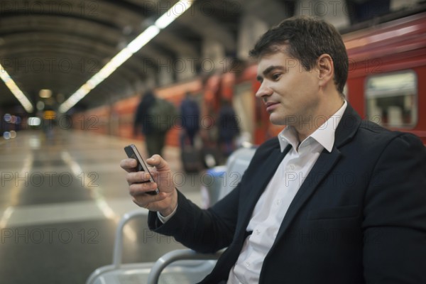 Young businessman using smartphone while waiting at the underground station. Blurred platform, train and people in background