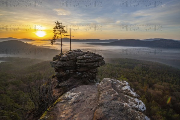 Sunrise on a rock with a summit cross and a view over a forest. Morning atmosphere with fog in the middle of nature. Landscape in the Palatinate Forest, Germany, Europe
