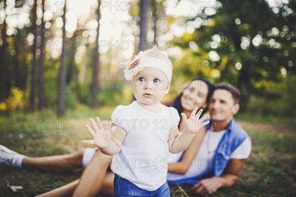 Little girl one year on the background of parents resting on graß learning to walk on nature in the park. The first steps of a child in nature outside the city in the forest