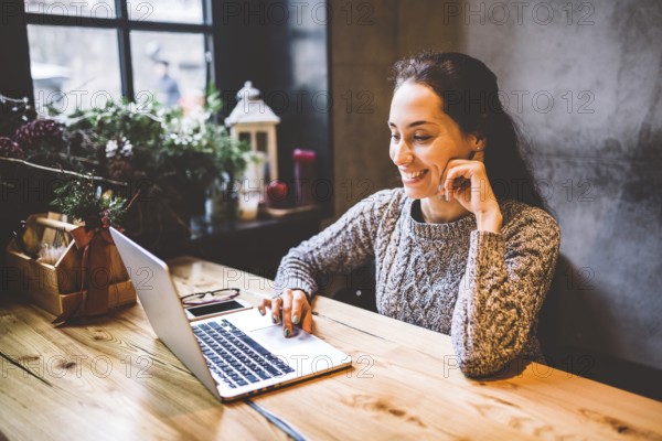 Beautiful young brunette woman using a laptop at coffee shop at a wooden table near window typing text on a keyboard. In winter, with the light from the lamp, she is dressed in a warm grey jumper