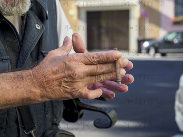 Older caucasian man sits on medical walker outdoors, smoking a cigar in public despite visible signs of frailty, illustrating contrast between age-related vulnerability and risky personal habits