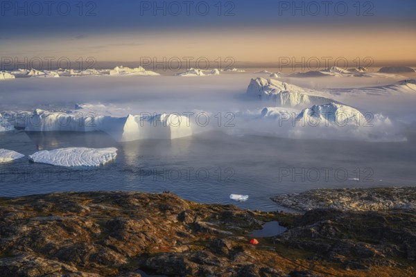 Large glaciers in the bay by the rocky shore in the light of the setting sun with fog over the water with orange tourist tent