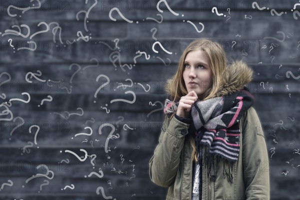 Pretty young female student in class with a problem to solve standing in front of a chalkboard in class covered in question marks looking thoughtfully into the air with her hand to her chin