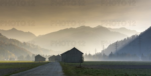 View of the Praettigau Valley near Klosters in Switzerland on an early morning with farm fields and wooden barns and power lines and mountains in silhouette behind in late autumn