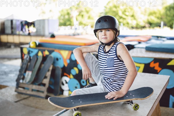 Boy sitting at skatepark, looking camera. Kid resting with skate board at skate park. child sits on ramp resting after training on skateboard. Trendy youngster enjoying free time at half pipe ramp