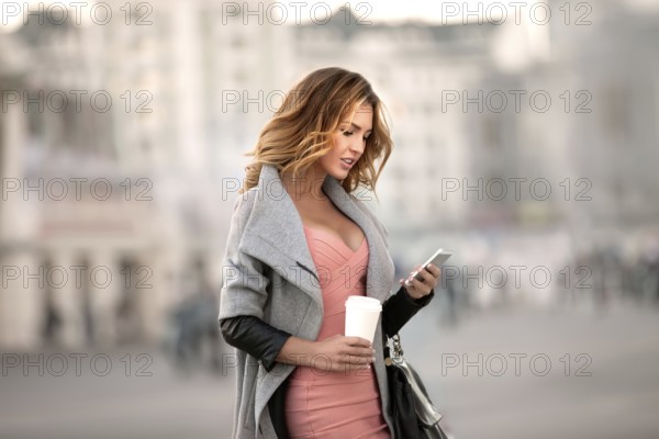 A businesswoman checking emails on her mobile phone and holding a coffee cup in her hand in front of an urban backdrop