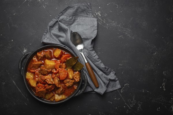 Beef meat stew with potatoes, carrot and delicious gravy in black casserole pot with bay leaves with spoon on black dark rustic concrete background from above, space for text, food photography, food photography