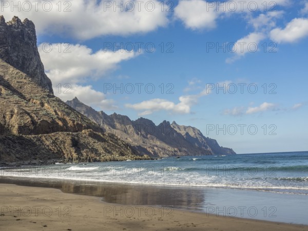 A picturesque beach with waves lapping against the sand and rocky cliffs and mountains in the background under a clear sky, tenerife, canary islands, spain