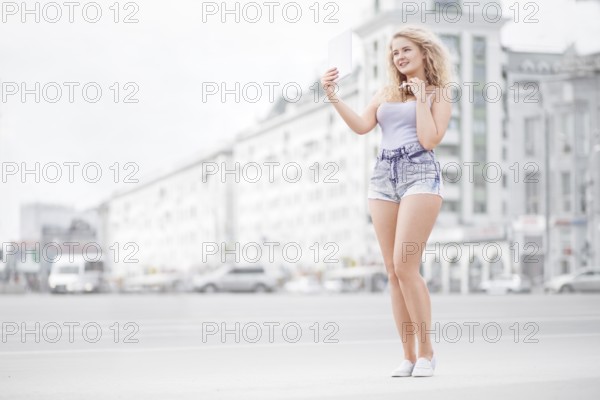 Happy young woman with vintage music headphones around neck taking selfie with tablet PC and smiling happily against urban city background