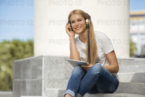 Happy young woman listening to music with vintage headphones, surfing the internet on a tablet PC and sitting on a staircase in front of an urban background
