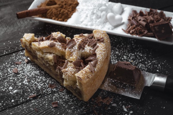 Slice of apple pie with powdered sugar and chocolate, on a knife, on a black rustic table and a plate with cinnamon.Sweet pastry. Baked desserts