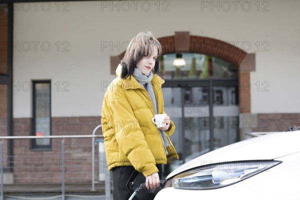 Woman in yellow jacket holding a cup, standing by charging electric car in urban environment, environmentally friendly