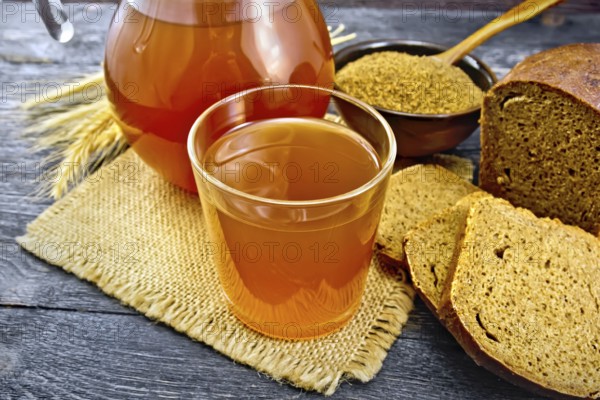 Kvass in glassful and glass jug on burlap, malt in a bowl, rye bread and ears on a wooden plank background