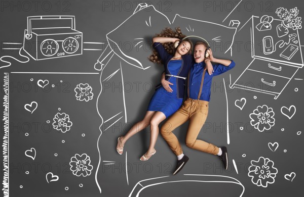 Happy valentines love story concept of a romantic couple lying in bed, sharing headphones, and listening to music against chalk drawings room background