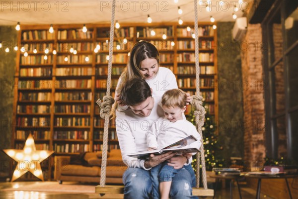 Family with baby relaxing at home swing seat in cosy dark living room on Christmas eve. Family having fun on the sea saw. Winter evening with family and child. Family reading book in living room