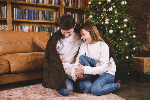 Happy family on Christmas Eve on the floor under a blanket near Christmas tree in the evening at home. Family spends Christmas time wrapped in rug during winter holidays. Winter magical time, fairytale vibes