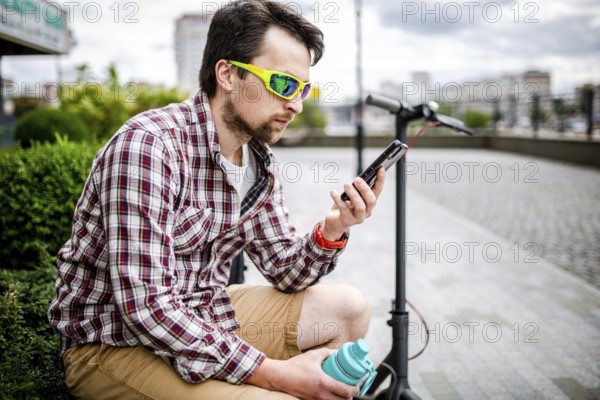 Caucasian man uses modern technologies Internet and cellular communication on smartphone while sitting on bench in city near electric scooter. Ecology and environmentally friendly urban transport