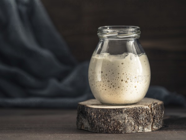 Active wheat sourdough starter in glass jar on brown wooden background. Starter for sourdough bread. Toned image. Copy space