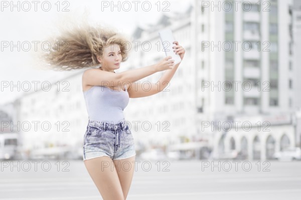 Happy young woman with vintage music headphones around neck taking selfie with tablet PC and smiling happily against urban city background