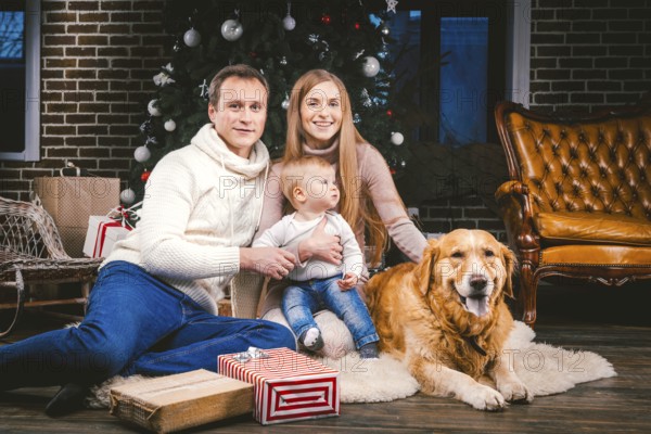 Theme Christmas and New Year family circle and domestic pet. Mom dad and child 1 year old Caucasian woman sitting on floor near Christmas tree with gifts and big dog breed labrador golden retriever