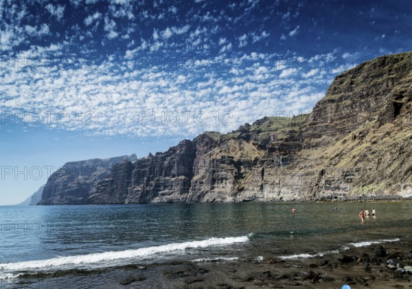 Tourists bathing at los gigantes volcanic black sand beach by famous natural cliffs landmark in south tenerife island spain