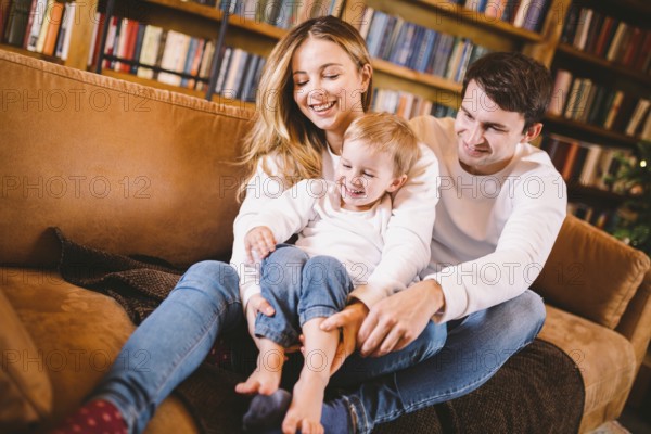 Christmas family portrait. Young Caucasian family in identical clothes having fun on sofa in living room with large library of books and Christmas tree in evening. Happy New Year and Merry Christmas