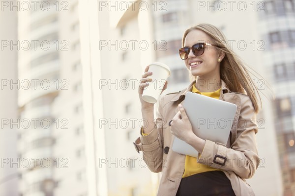 Beautiful young businesswoman with a disposable coffee cup, drinking coffee and holding a tablet in her hands, against an urban background