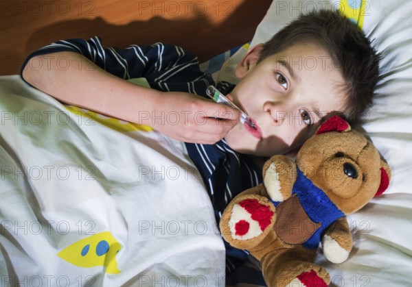 Sick child in bed with teddy bear. Measuring the temperature with a thermometer