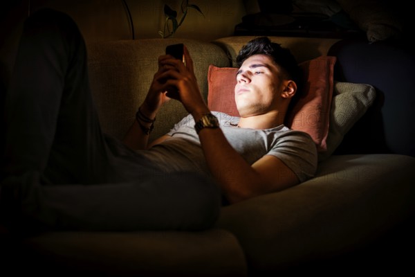 Attractive Young Man Lying on Sofa at Night and Illuminated by Light from Screen of Cell Phone at Home in Living Room