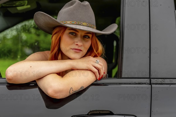 A beautiful redhead model sits in her truck while wearing a cowboy hat on her farm