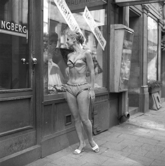In the 1950s. A young woman dressed in underwear. She is wearing a bra and panties and is standing outside a shop looking into the shop window where signs are posted with the text Sale Discounts Sweden 1959. Kristoffersson ref CG20-11
