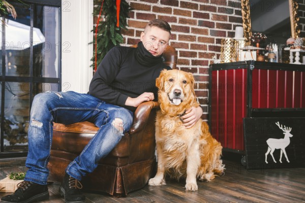 The theme is the friendship of man and animal. Caucasian young male and pet dog breed Labrador Golden Retriever at home inside in winter. Christmas time, New Year holidays