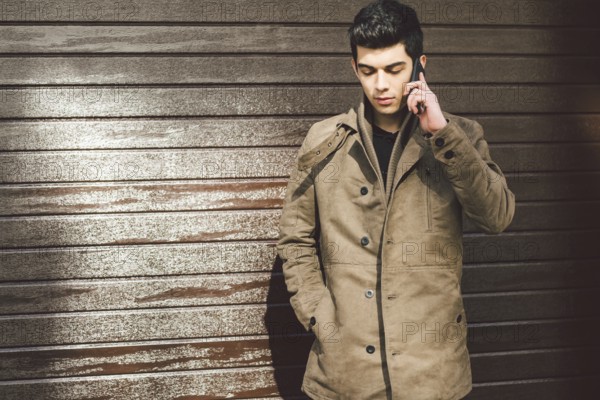 Portrait of a model young mulatto male Turkish Mediterranean race brunet in coat uses hand phone technology businessman on the street in sunny weather against the background of a wooden wall