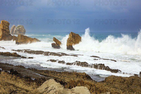 Storm waves break on the coastal rocks. Snow-white sea foam glistens in the dim autumn sun. Cantabria, Spain. Playa de la Arnia beach. Amazing geological formations amaze tourists