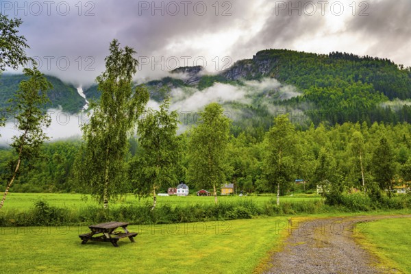 Rain-wet dirt road crosses a valley. Jostedalsbreen Park. Heavy clouds covered the sky. Valley surrounded by high forested mountains. Cold summer in Norway