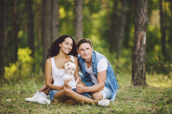 The theme family outdoor recreation in forest park area. Young beautiful stylish caucasian family with small child daughter one year old sit on the graß in the coniferous forest in summer at sunset