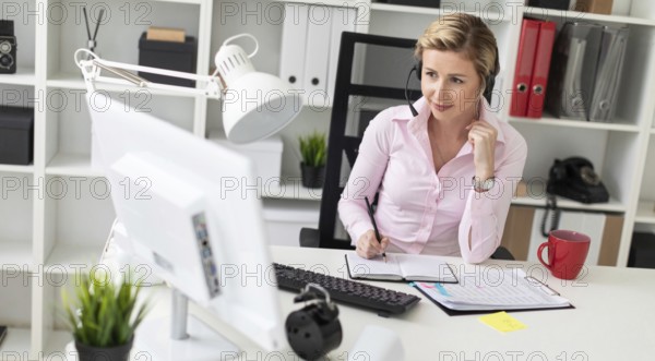 A young girl in headphones sits at a table in the office, holds a pencil in her hand and works with a notebook and a computer