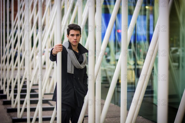 Stylish Young Handsome Man in Black Coat Standing in City Centre Street among White Metal Bars of Moden Architecture