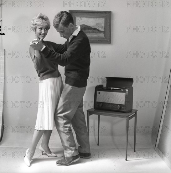Learning to dance in the 1950s. A young copule is dancing in a photographers studio. The young woman seems to be the one who knows how to dance. Her boyfriend does not seem to know the steps and is struggling with the dance moves. To create the mood of an at home environment a painting is put on the wall and a combined radio and gramophone is visible. Sweden 1959. Kristoffersson ref CG36