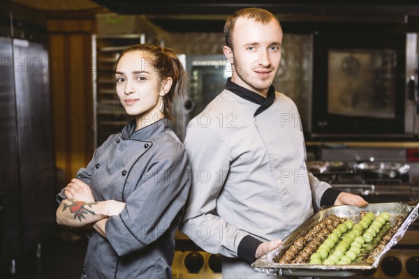 Profession preparation confectionery products. Caucasian man and woman two employees colleague teamwork pastry chef posing looking camera in uniform in kitchen holding sweets dessert candy truffle