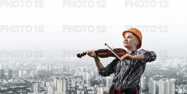 Beautiful young woman in safety helmet playing violin. Smiling professional female builder in checkered blue shirt on cityscape background. Industrial architecture and construction concept