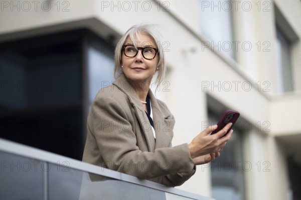 A senior businesswoman stands outside a contemporary building, holding her smartphone. She appears thoughtful and engaged, soaking in the sunlight