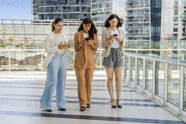 Three businesswomen are walking while using smartphones outdoors. All women are dressed in business attire