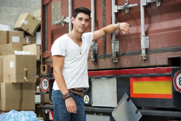 Waist Up Portrait of Young Smiling Man Next to Freight Truck Leaning against it, Looking at Camera