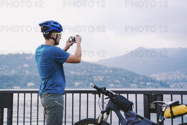 Cyclist taking pictures with smart phone. Caucasian man in bike helmet taking photo of landscape. Outdoor activity. Tourist makes photo of Lake Como in Italy. travel people lifestyle