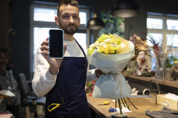 Ein florist mit einem strauß in den händen zeigt den bildschirm eines smartphones vor dem hintergrund eines blumenladens